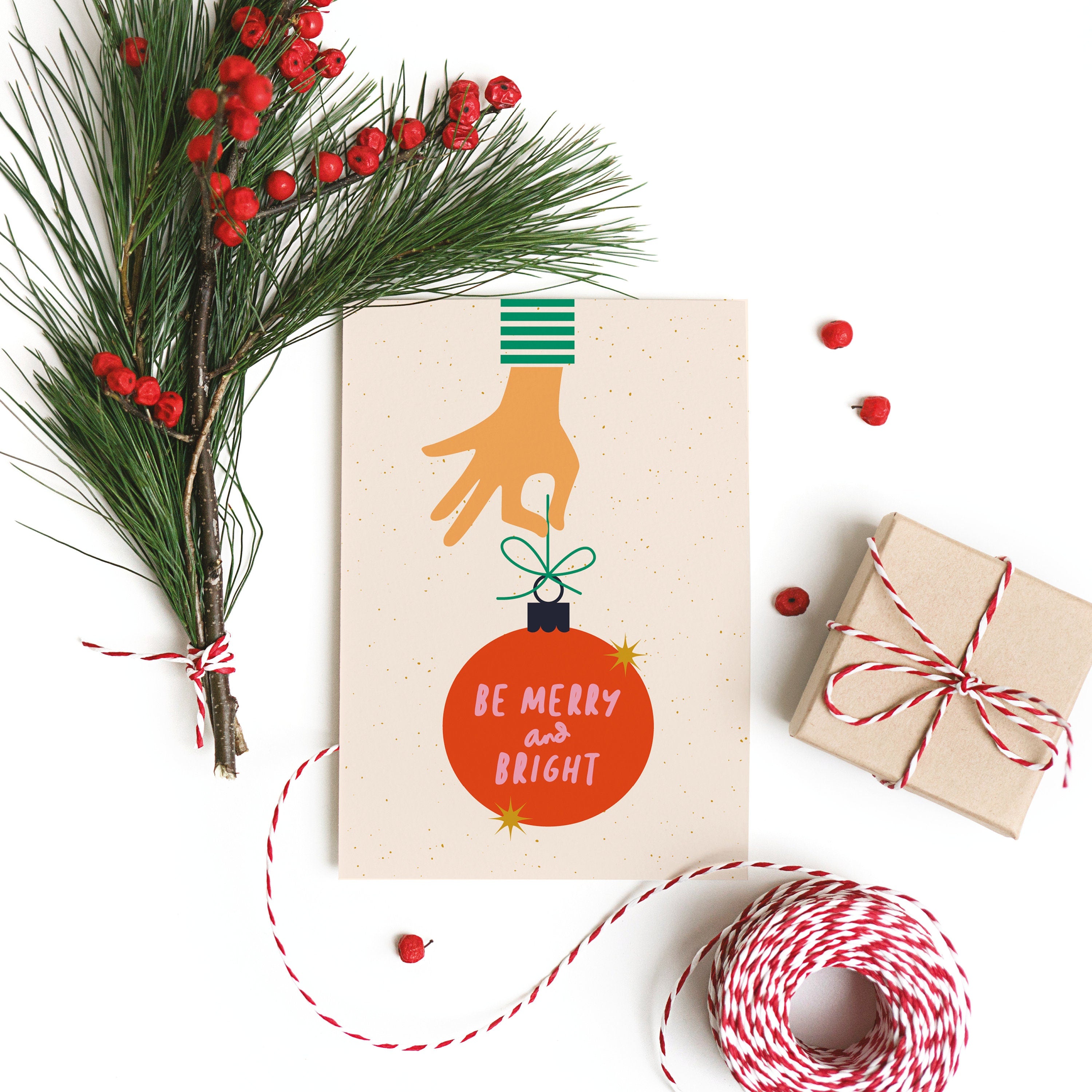 christmas card with a hand holding red christmas tree ornament and a text in it that says be merry and bright